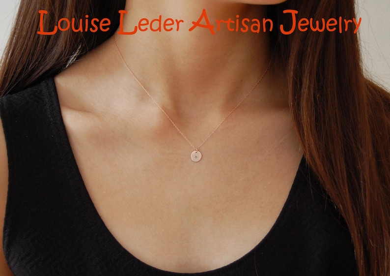 May include: A delicate rose gold necklace featuring a small, circular pendant with a single engraved letter. The text Louise Leder Artisan Jewelry is displayed in orange at the top of the image. The necklace rests against a black sleeveless top.