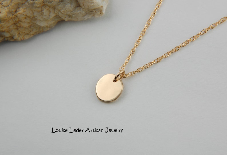 May include: A gold pendant necklace featuring a small, circular charm. The fine chain and charm are set against a plain backdrop. The text "Louise Leder Artisan Jewelry" is visible.