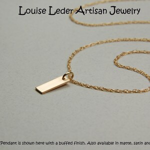 May include: A gold-colored necklace with a rectangular pendant. The chain is delicate and the pendant is a simple, polished rectangle. The text "Louise Leder Artisan Jewelry" is at the top. The text at the bottom says "Pendant is shown here with a buffed finish. Also available in matte, satin and shiny."