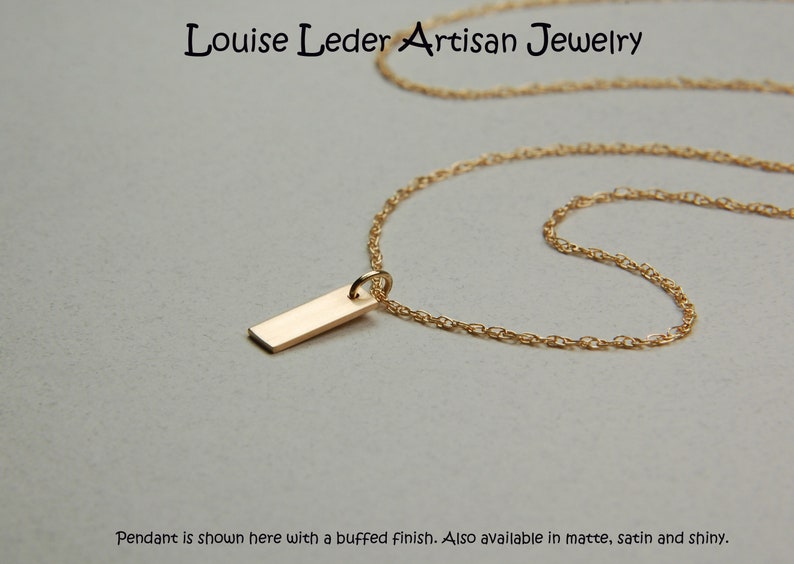 May include: A gold-colored necklace with a rectangular pendant. The chain is delicate and the pendant is a simple, polished rectangle. The text "Louise Leder Artisan Jewelry" is at the top. The text at the bottom says "Pendant is shown here with a buffed finish. Also available in matte, satin and shiny."