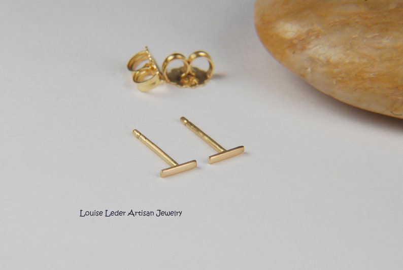 May include: A pair of gold bar stud earrings with matching earring backs. The earrings are small, minimalist, and have a simple, modern design. The earrings are displayed on a white surface.