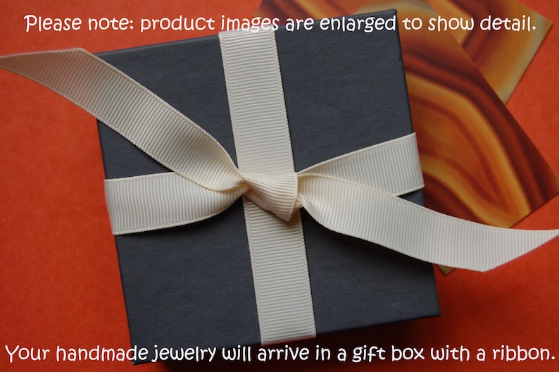 May include: A dark grey gift box tied with a cream ribbon, set against an orange backdrop. The text at the top reads, "Please note: product images are enlarged to show detail." The text at the bottom says, "Your handmade jewellery will arrive in a gift box with a ribbon."
