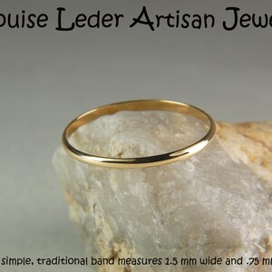 May include: A gold-coloured ring band resting on a textured stone. The text at the top reads Louise Leder Artisan Jewelry, and the text at the bottom reads This simple, traditional band measures 1.5 mm wide and .75 mm thick. The ring is smooth.