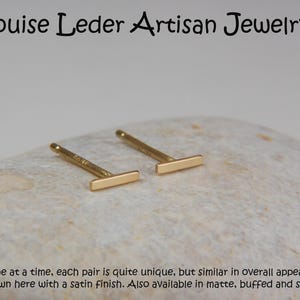 May include: A pair of gold-colored bar stud earrings. The earrings are displayed on a light-colored, textured surface. The text "Louise Leder Artisan Jewelry" is at the top. The text at the bottom says the earrings are unique.