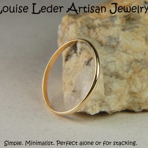 May include: A thin, polished gold band ring with a smooth, rounded surface. The text 'Louise Leder Artisan Jewelry' appears at the top, and the text 'Simple. Minimalist. Perfect alone or for stacking.' appears at the bottom of the image.