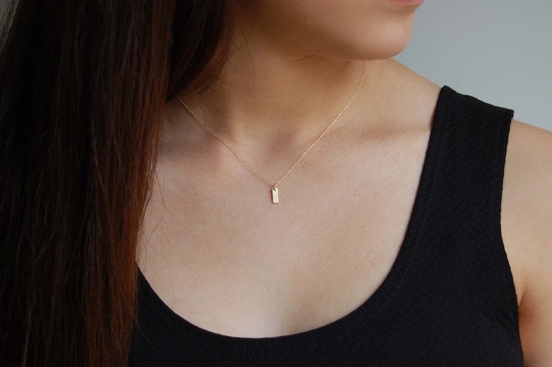 May include: A delicate gold necklace with a small, rectangular pendant. The pendant hangs from a fine chain, creating a minimalist and elegant look. The necklace is shown against a dark background.