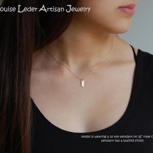 May include: A delicate gold necklace featuring a small, rectangular pendant. The pendant, approximately 10 mm in size, hangs from a thin chain. The jewelry is from Louise Leder Artisan Jewelry.