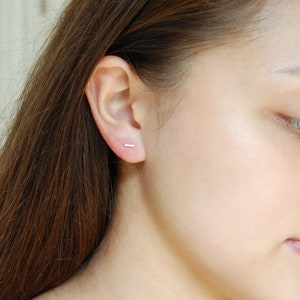 May include: A close-up of a person's ear with a small, silver bar earring. The earring is simple and understated, and it is a popular choice for minimalist jewelry.