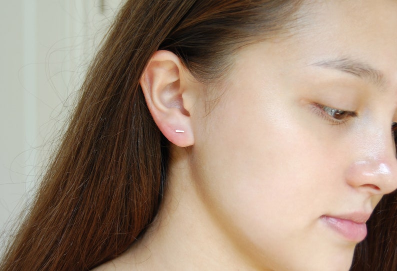 May include: A close-up of a person's ear with a small, silver bar earring. The earring is simple and understated, and it is a popular choice for minimalist jewelry.
