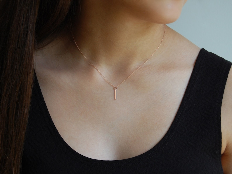 Puede incluir: Un delicado collar de oro rosa con un colgante de barra vertical y delgada. El colgante cuelga de una fina cadena, creando un dise&ntilde;o minimalista. El collar se muestra sobre una camiseta negra.