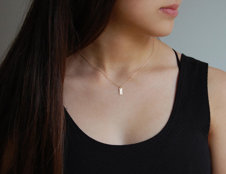 May include: A delicate gold necklace featuring a small, rectangular pendant. The pendant hangs from a fine chain, creating a minimalist and elegant look. The necklace is worn against a black top, highlighting its subtle shine.