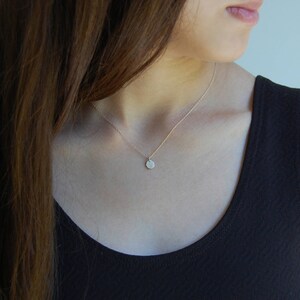 May include: A fine gold necklace featuring a small, circular pendant. The pendant has a textured surface. The necklace is worn over a black top, with long brown hair in the background.