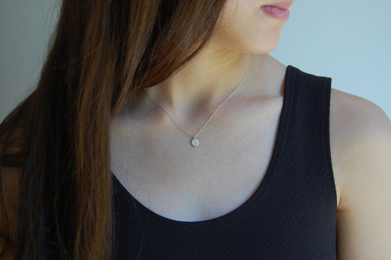 May include: A fine gold necklace featuring a small, circular pendant. The pendant has a textured surface. The necklace is worn over a black top, with long brown hair in the background.