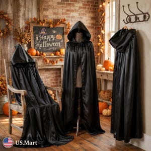 May include: Three black hooded capes made of a smooth, satin-like fabric. One cape is draped over a wooden chair, one is displayed on a dress form, and one hangs from a metal wall hook. Each cape features a deep hood and long, flowing fabric.