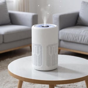May include: A white air purifier with a cylindrical design, emitting a vapor. The device has a perforated pattern on the sides and sits on a round, white table. The background features two gray sofas.