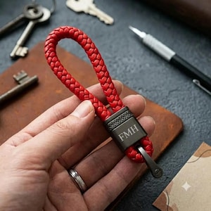 May include: A red braided leather keyring featuring a dark gunmetal grey rectangular metal clasp engraved with the letters FMH. The braided loop is attached to the metal hardware, which includes a small circular ring at the bottom end.
