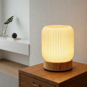 Puede incluir: Lámpara de mesa cilíndrica con pantalla acanalada blanquecina y base de madera. La lámpara está iluminada, emitiendo un brillo cálido. Está sobre una mesita de noche de madera. Dimensiones: aproximadamente 15 cm de diámetro y 20 cm de alto.
