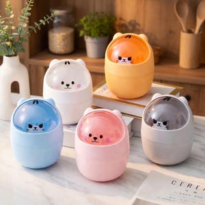 Puede incluir: Un conjunto de cinco mini papeleras con forma de oso en colores vibrantes: naranja, blanco, rosa, azul y gris. Cada papelera tiene una tapa transparente y redondeada y una cara de oso de dibujos animados.