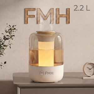 May include: A 2.2 L humidifier with a cylindrical, transparent amber-tinted water tank and a white base. The device features a central glowing light, a wood-toned top lid, and the text FMH printed on the front of the base and on the wall behind.