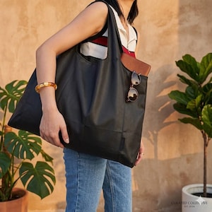 May include: A black tote bag with a rectangular shape. The bag has a pocket on the front. Sunglasses and a brown wallet are visible inside the bag. A gold-coloured bracelet is on the person's wrist. The bag is being held.