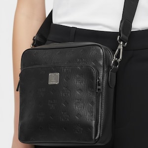 May include: Black crossbody bag with a repeating embossed pattern. The bag has a rectangular shape with a front pocket and a silver-tone metal plate. The adjustable strap is black with white text. The bag is made of a textured material.