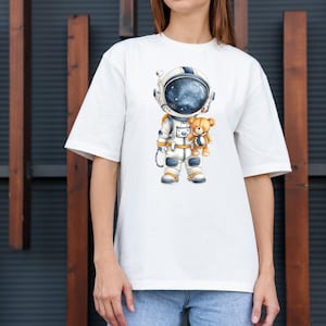 May include: White t-shirt featuring a cartoon astronaut in a spacesuit holding a teddy bear. The astronaut's helmet shows a starry galaxy. The astronaut's suit is white and blue with gold accents. The teddy bear is light brown.