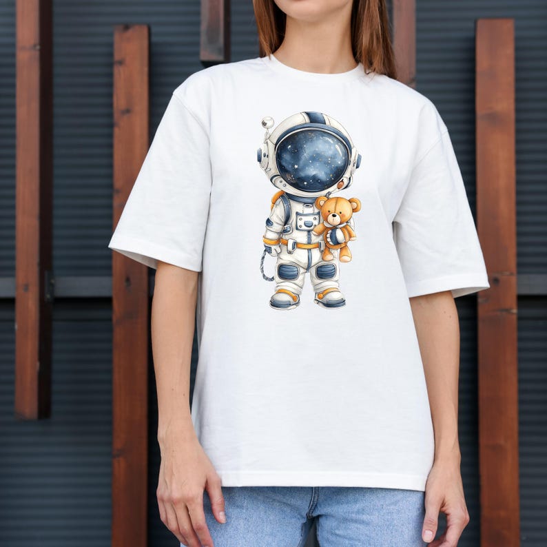 May include: White t-shirt featuring a cartoon astronaut in a spacesuit holding a teddy bear. The astronaut's helmet shows a starry galaxy. The astronaut's suit is white and blue with gold accents. The teddy bear is light brown.
