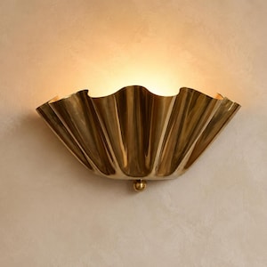 May include: A semi-circular wall sconce crafted from polished brass with a distinct scalloped, pleated design. The fixture features a small spherical brass finial at the base and emits a warm, soft glow from the top, casting light upward.
