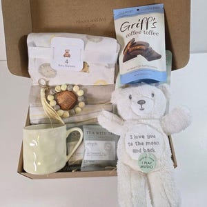 May include: A gift box containing a set of four baby blankets with lion prints, a bag of Griff's coffee toffee, a beaded keychain, a light yellow textured mug, tea bags, and a white plush teddy bear with the text I love you to the moon and back.