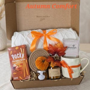 May include: A cardboard gift box labeled Autumn Comfort containing a white knit blanket tied with an orange ribbon, a box of Pocky Pumpkin Spice, a small orange fabric pumpkin, a round coaster, a jar of Rosemary candle, and a white mug.