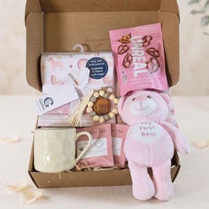 May include: A gift box containing a pink Gerber blanket with rainbow patterns, a bag of Unreal milk chocolate covered pretzels, a light-colored textured ceramic mug, a wooden bead keychain with the word MAMA, and a pink plush bear with My First Bear text.