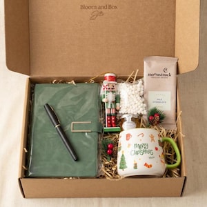 May include: A cardboard gift box labeled Bloom and Box containing a green notebook with a black pen, a nutcracker figurine, a bag of white marshmallows, a small jar, a Moonstruck milk chocolate bar, and a white mug with Christmas graphics.