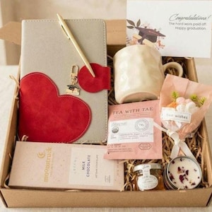May include: A gift box containing a gray notebook with a red heart-shaped cover, a gold pen, a graduation card, a textured cream mug, a Tea With Tae tea bag, a Moonstruck milk chocolate bar, a small jar of honey, and a floral bouquet with a tag.