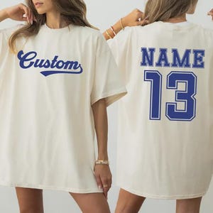 May include: Cream-colored t-shirt with the word "Custom" in a navy blue script on the front. The back of the shirt features the word "NAME" above the number "13" in navy blue block letters.