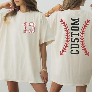 May include: Cream-colored t-shirt with a baseball-themed design. The front features the number "13" in red, with a baseball stitch pattern. The back displays the word "CUSTOM" in black, framed by red baseball stitch lines.