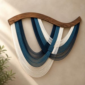 Op de afbeelding: Een wandhanger met een gebogen, donkerbruin houten bovendeel en meerdere strengen wit, lichtblauw en donkerblauw koord die in overlappende, U-vormige lussen zijn gedrapeerd, wat een gelaagd en symmetrisch patroon op het oppervlak creëert.