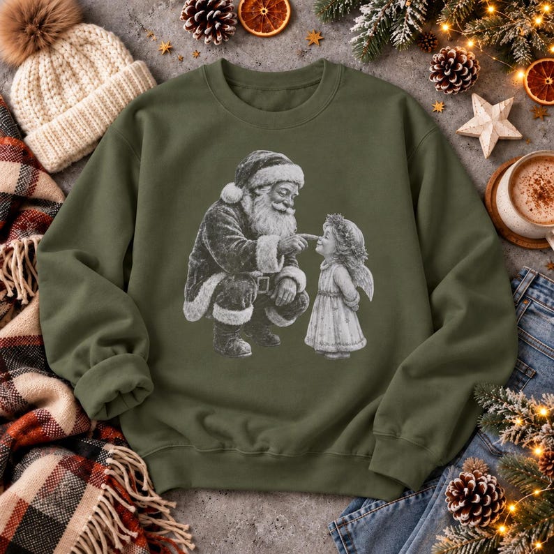Olivgrünes Weihnachts-Sweatshirt in Military Green: Weihnachtsmann berührt die Nase des Christkindls. Flatlay auf Beton mit umgeschlagenem Ärmel und Weihnachtsdeko.