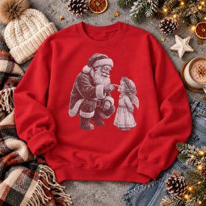 Rotes Weihnachts-Sweatshirt mit nostalgischem Weihnachtsmann-und-Christkindl-Motiv und leicht transparenter Druckoptik. Flatlay auf Beton, linker Ärmel umgeschlagen.