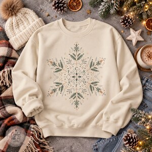 Könnte beinhalten: Ein cremefarbenes Sweatshirt mit Rundhalsausschnitt und einem zentralen Schneeflocken-Motiv aus grünen Tannenzweigen, braunen Tannenzapfen und kleinen Sternen. Die Buchstaben F, C, K, A und D sind kreuzförmig im Schneeflocken-Design angeordnet.
