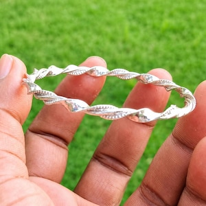 Puede incluir: Una pulsera tipo brazalete de color plateado con un diseño de cuerda trenzada continua. La superficie metálica tiene un acabado pulido con segmentos alternos lisos y texturizados que crean un patrón de espiral repetitivo alrededor de la banda.