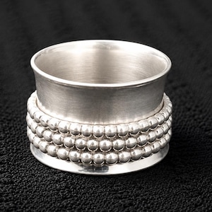Peut inclure: Un rond de serviette en métal argenté doté d'une large bande centrale lisse, entourée de trois rangées de petites perles sphériques sur l'extérieur. L'objet présente une finition polie et réfléchissante ainsi qu'une forme cylindrique.