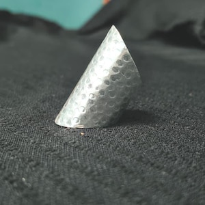 May include: A silver-colored metal ring with a wide, slanted, and tapered band design. The surface of the ring features a distinct hammered texture, creating a series of small, circular indentations across the entire exterior of the metal band.