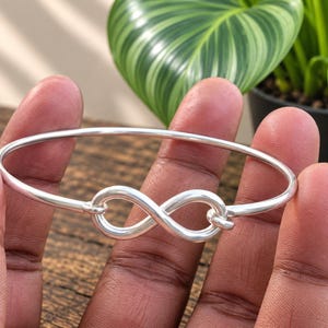 Może przedstawiać: Polerowana bransoletka typu bangle w kolorze srebrnym z centralnym motywem symbolu nieskończoności. Bransoletka wykonana jest z gładkiego, ciągłego metalowego drutu, który z przodu tworzy pętlę w kształcie ósemki, nadając jej prosty wygląd.