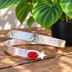 Pode incluir: Uma pulseira de enrolar em tom prateado com uma textura martelada na banda superior e um acabamento liso na banda inferior. A extremidade da pulseira é acentuada com uma única pedra vermelha oval e um pequeno design de estrela de cinco pontas.