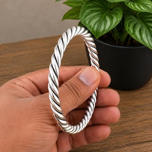 Puede incluir: Un brazalete rígido de tono plateado con un diseño continuo de cuerda trenzada. La superficie metálica es pulida y reflectante, mostrando un patrón de espiral uniforme que envuelve toda la circunferencia de la pieza de joyería circular.