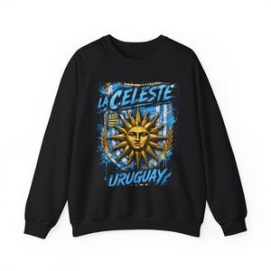 May include: Black sweatshirt featuring a graphic design with the text "LA CELESTE" and "URUGUAY". The design includes a sun with a face, blue and white stripes, and a splash effect.