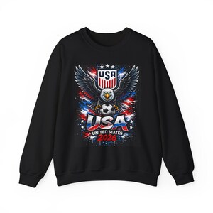 May include: Black sweatshirt featuring a graphic of an eagle with outstretched wings, a soccer ball, and the text "USA UNITED STATES 2026." The design incorporates red, white, and blue elements, with the USA logo at the top.