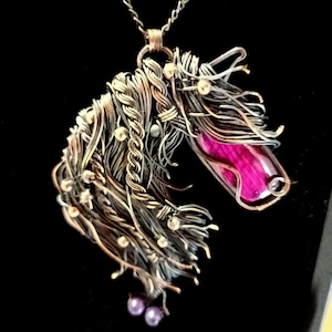 Puede incluir: Un collar con colgante hecho a mano con un diseño de cabeza de caballo. El colgante incorpora una gema rosa vibrante y está adornado con pequeños acentos brillantes. La melena del caballo está intrincadamente tejida con alambre de plata y cobre, y el collar tiene una cadena de plata.