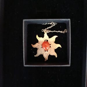 Puede incluir: Un collar con colgante dorado con un diseño de sol y una piedra central de color rojo anaranjado. El colgante se muestra en una caja negra, resaltando los detalles y el contraste de color de la joya.