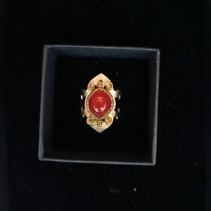 Puede incluir: Un anillo dorado con una piedra preciosa roja ovalada en el centro. El anillo tiene un diseño ornamentado con detalles de filigrana alrededor de la piedra preciosa. El anillo se presenta en una caja negra.